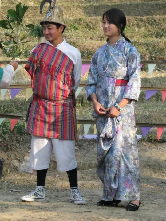 पारंपरिक भूटानी या हिमालयी वेशभूषा में एक युवा लेप्चा पुरुष और महिला एक बाहरी सेटिंग में खड़े हैं, पुरुष ने धारीदार गाउन और टोपी पहनी है, जबकि महिला ने नीले रंग की किमोनो-शैली की पोशाक और हार पहना है।